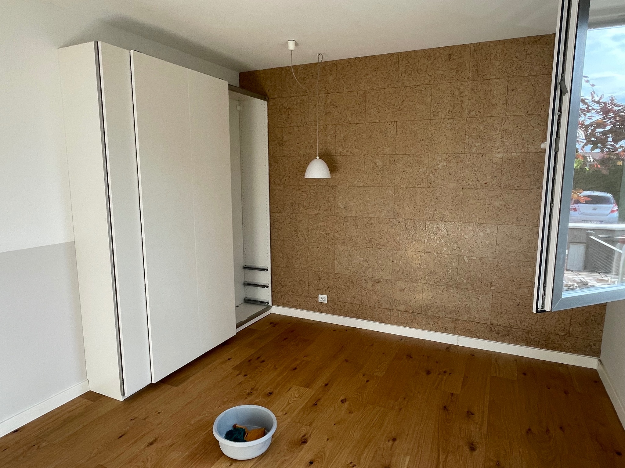 Pokój po remoncie: drewniana podłoga, ściana wyłożona korkiem, biała szafa w zabudowie, wisząca lampa i otwarte okno z widokiem na zewnątrz; na podłodze wiadro z narzędziami.