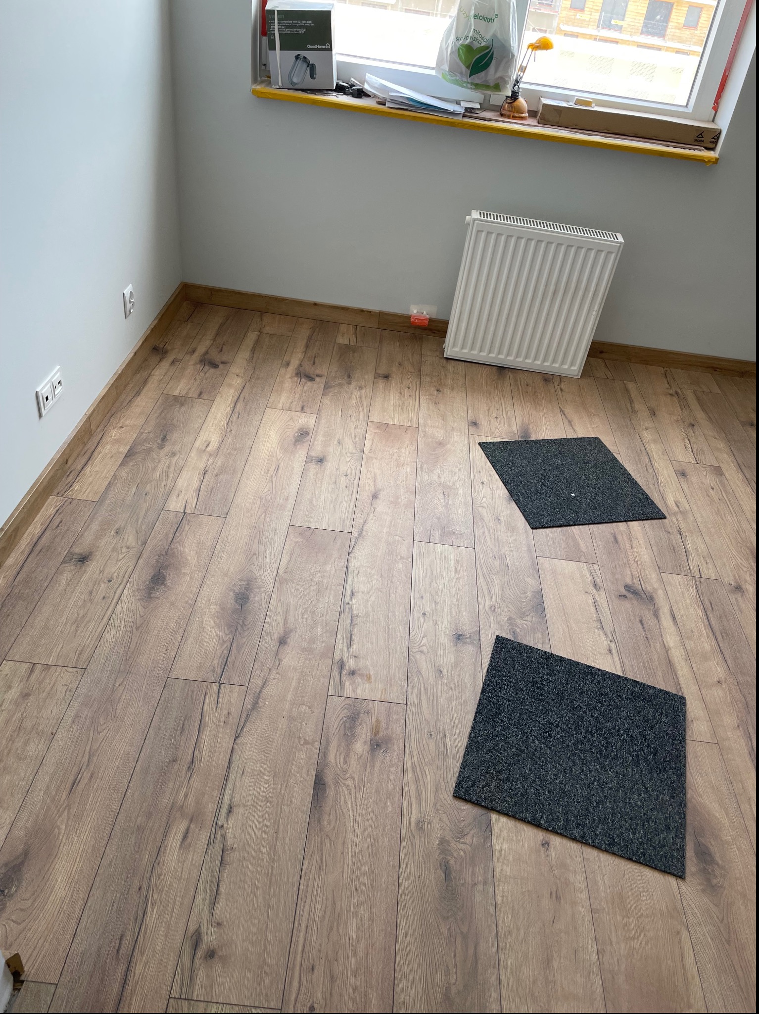 Wykończone wnętrze pokoju z panelami podłogowymi o wzorze drewna, białymi ścianami, grzejnikiem pod oknem i dwoma szarymi dywanikami.