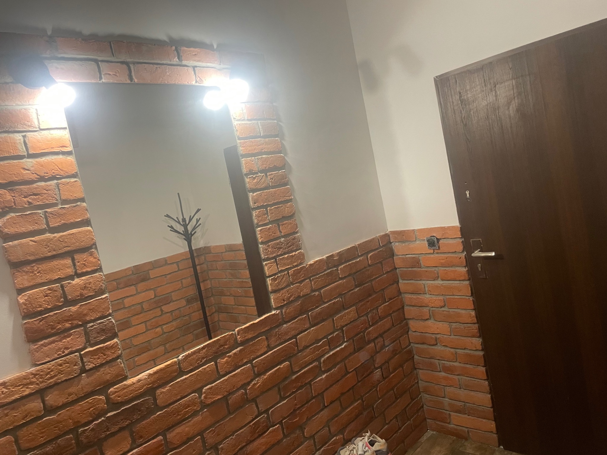Fragment korytarza z lustrem oprawionym w cegłę klinkierową, oświetlonym punktowymi lampami, obok ciemne drzwi.