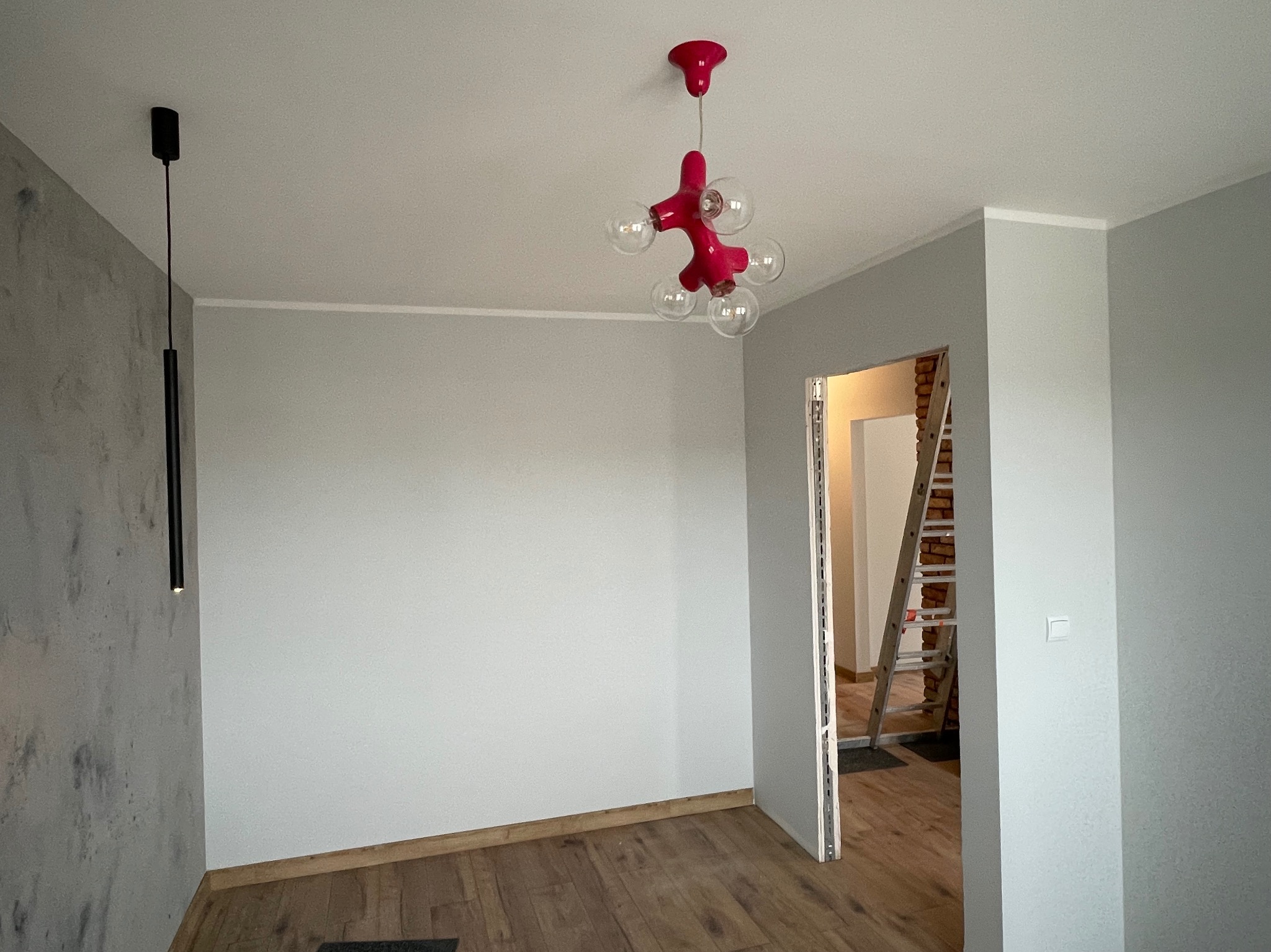 Wnętrze pokoju w trakcie remontu z szarą ścianą z efektem betonu, drewnianą podłogą, oryginalną czerwoną lampą sufitową i widoczną drabiną w przejściu do drugiego pomieszczenia.