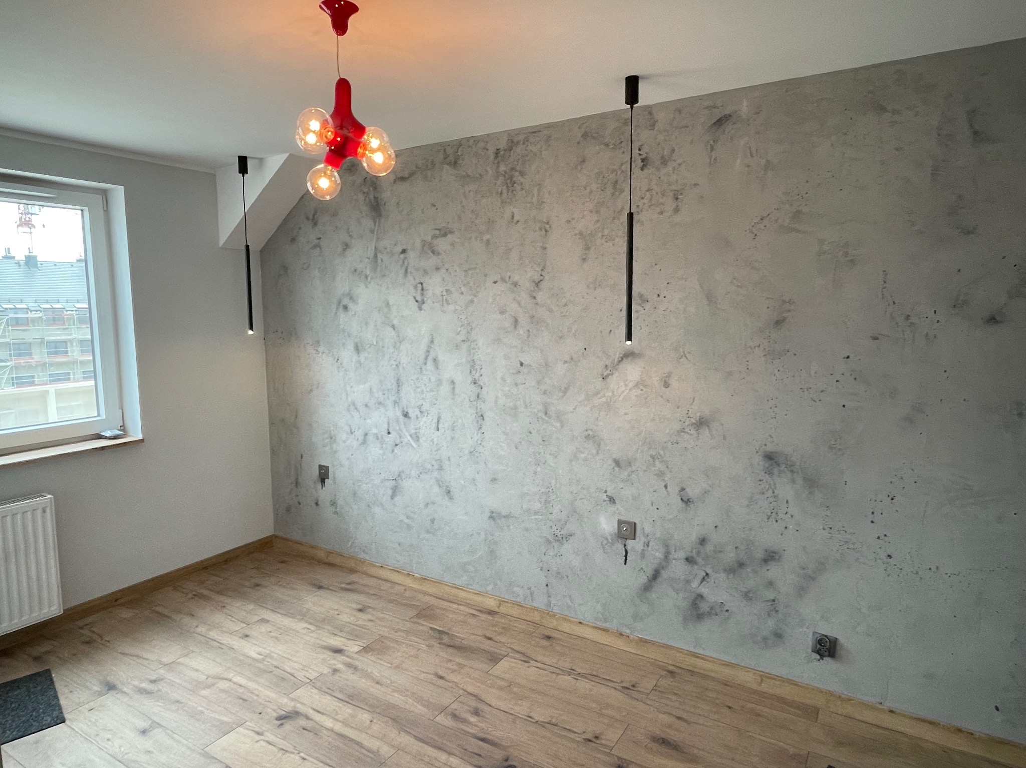 Pokój po zakończonym remoncie, z parkietem, ścianą pokrytą dekoracyjną szarą strukturą i nowoczesnym oświetleniem.