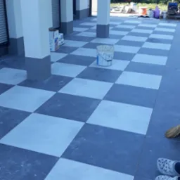 Szachownica z jasnych i ciemnych płytek na tarasie, widoczny kubeł z farbą i fragment butów w rogu.