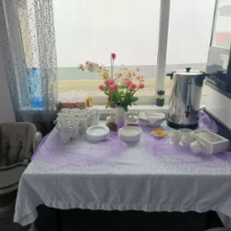Stół z białym obrusem i fioletową dekoracją, zastawiony filiżankami, talerzami, kwiatami i podgrzewaczem wody. Obok krzesełko do karmienia dziecka.