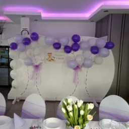 Dekoracja sali na Pierwszą Komunię Świętą: biało-fioletowe balony, ścianka dekoracyjna z motywem liści, kwiaty i elegancko nakryty stół. Fioletowe akcenty na krzesłach.