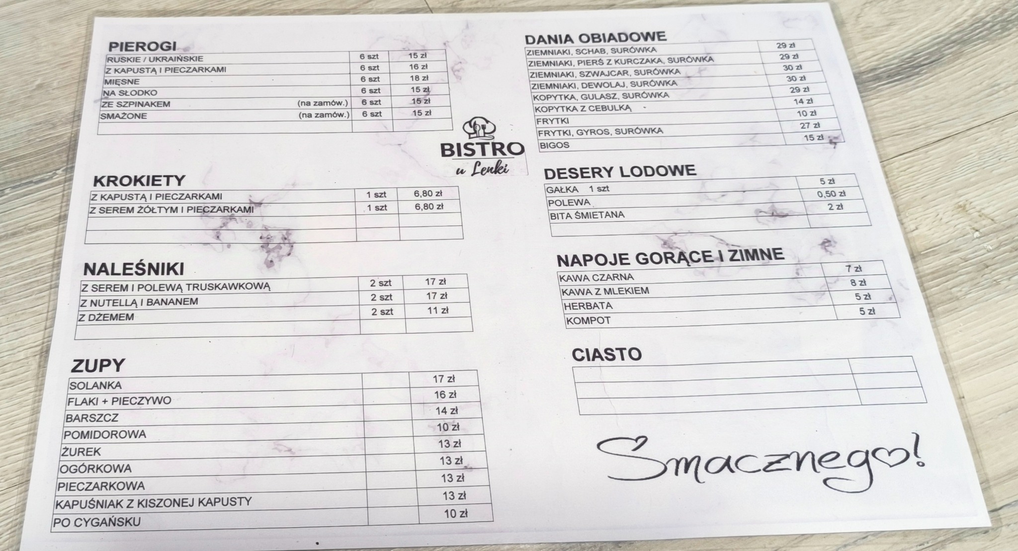 Karta menu Bistro u Lenki z ofertą dań obiadowych, pierogów, krokietów, naleśników, zup, deserów lodowych i napojów, na tle drewnianego stołu, z dopiskiem 'Smacznego!' na dole.