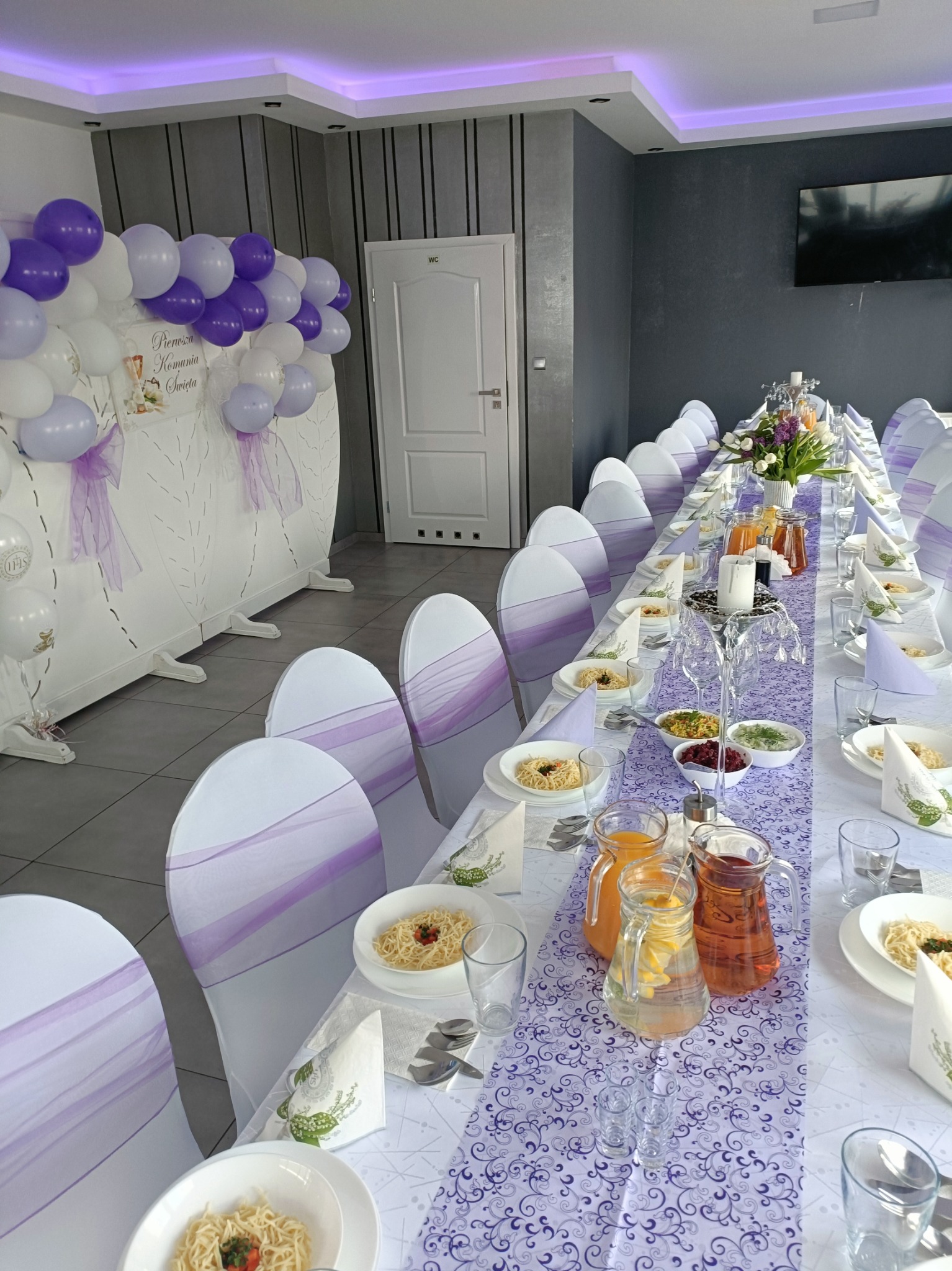Uroczysty, długi stół z białym obrusem w fioletowe wzory, dekoracje z białych i fioletowych balonów. Krzesła z fioletowymi szarfami.