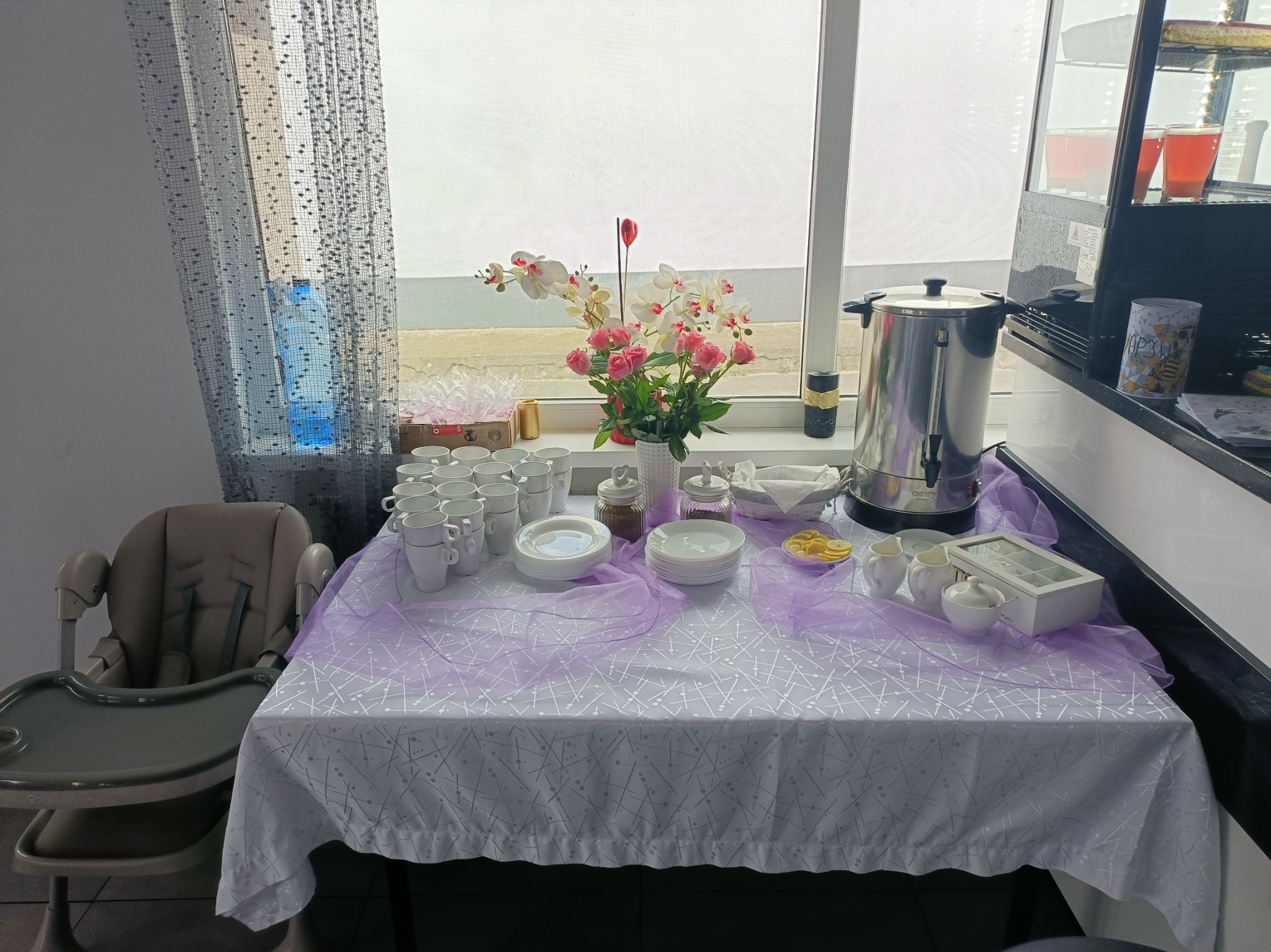 Stół z białym obrusem i fioletową dekoracją, zastawiony filiżankami, talerzami, kwiatami i podgrzewaczem wody. Obok krzesełko do karmienia dziecka.