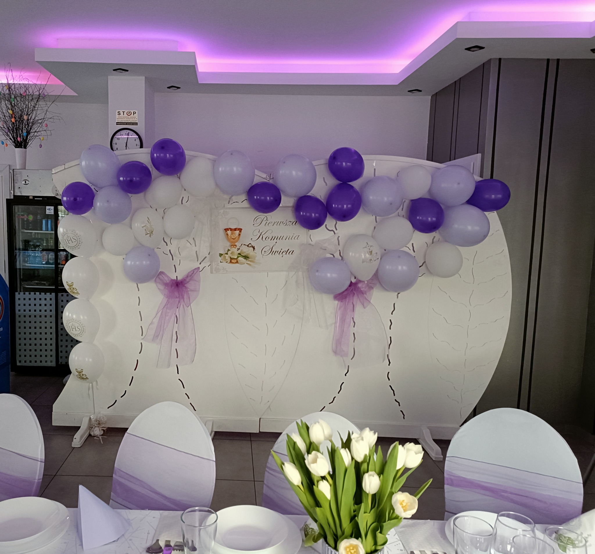 Dekoracja sali na Pierwszą Komunię Świętą: biało-fioletowe balony, ścianka dekoracyjna z motywem liści, kwiaty i elegancko nakryty stół. Fioletowe akcenty na krzesłach.