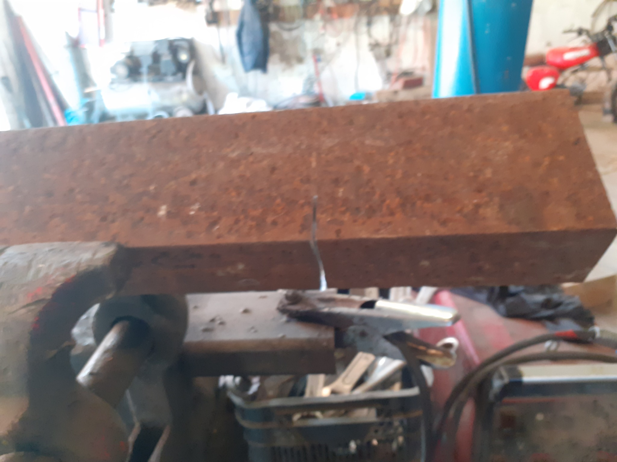 Zardzewiały, prostokątny kawałek metalu zamocowany w starym imadle, z widocznym śladem cięcia i narzędziami w tle warsztatu.