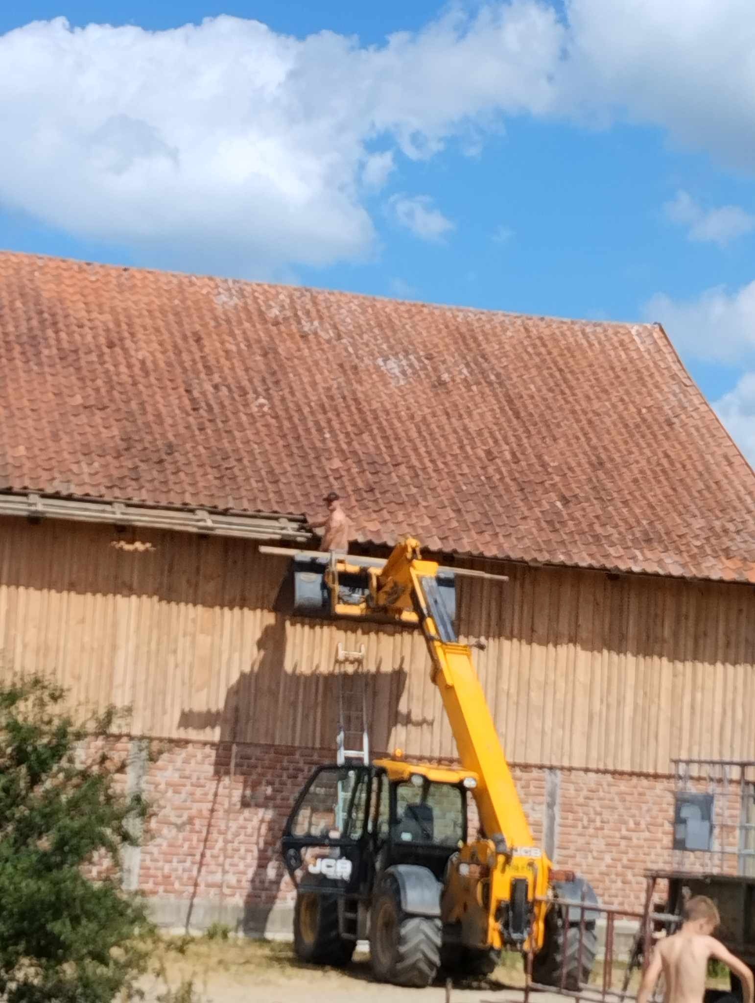 Pracownik na podnośniku teleskopowym, na tle drewnianego budynku z czerwoną dachówką, wykonuje prace na wysokości, widoczne niebo z chmurami.