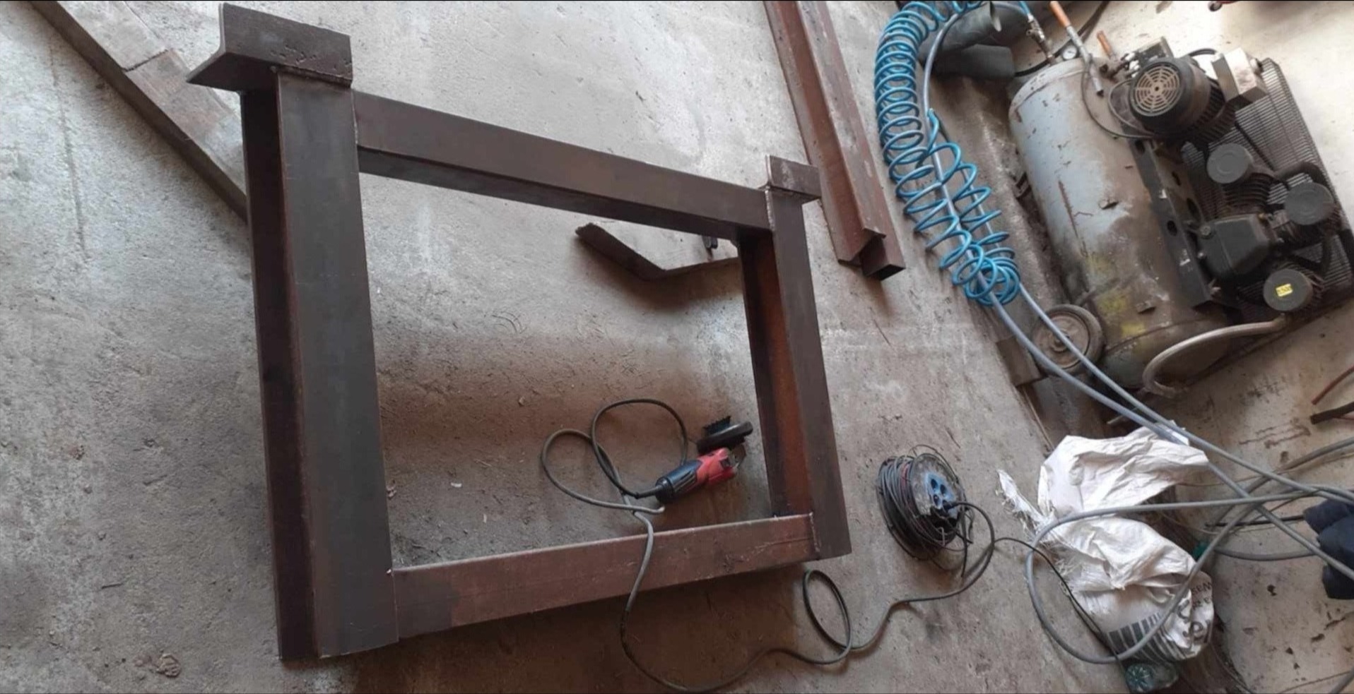 Metalowa rama spawana z profili stalowych, szlifierka kątowa obok, w tle sprężarka powietrza i przewody na betonowej posadzce.