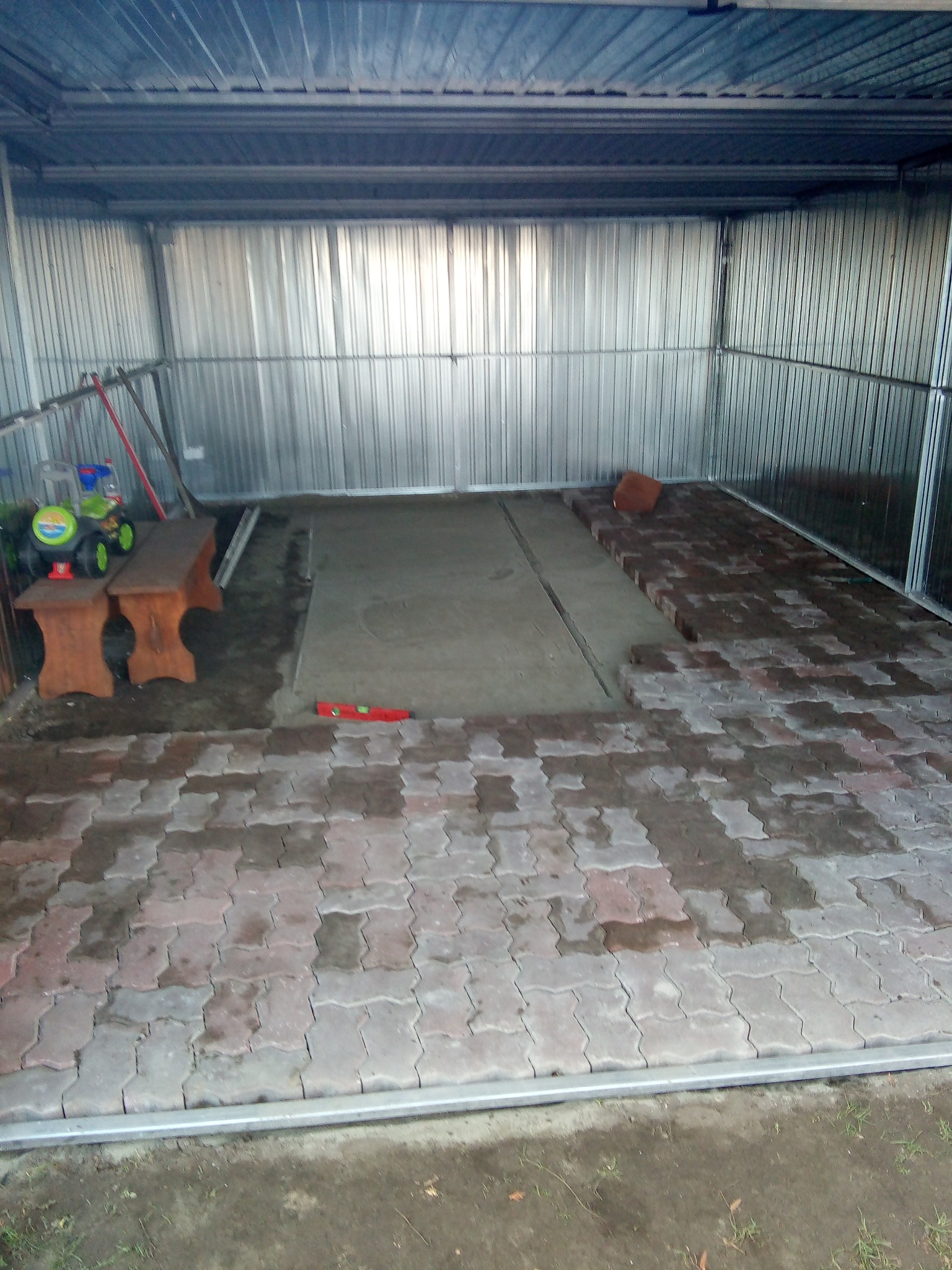 Wnętrze metalowego garażu z częściowo ułożoną kostką brukową, widoczny poziomica, dwie drewniane ławki i zabawki dziecięce w rogu.