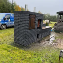 Murowany piec zewnętrzny z ciemnej cegły z drewnianymi drzwiczkami i dwoma kominami, zrealizowany w Suwałkach. W tle widoczny niebieski samochód dostawczy i mieszalnik do betonu.