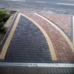 Ułożona kostka brukowa w odcieniach szarości i czerwieni, wzbogacona o dekoracyjne pasy w kolorze beżowym, zakończona odpływem liniowym.