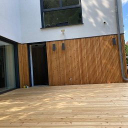 FachBud Rafał Szkwerko - Nowoczesny taras z jasnego drewna, zintegrowany z elewacją budynku wykończoną drewnianymi panelami. Widoczne okno i drzwi tarasowe z czarnymi ramami.