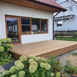 FachBud Rafał Szkwerko - Jasny, drewniany taras przylegający do białego domu z brązowymi oknami, otoczony zielenią i kwiatami hortensji. Widoczny fragment sąsiedniego domu.