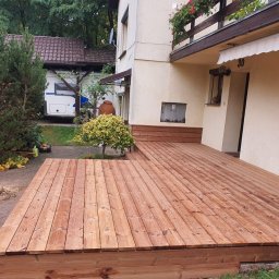 FachBud Rafał Szkwerko - Nowy, drewniany taras przydomowy z desek w ciepłym odcieniu brązu, z niskim stopniem i roślinnością w tle. Widoczny fragment elewacji budynku z numerem 30.