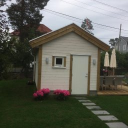 FachBud Rafał Szkwerko - Mała, biała altana z drewnianymi detalami, oświetlona lampami ściennymi, otoczona zielonym trawnikiem i ścieżką z płyt. W tle widoczny taras i meble ogrodowe.