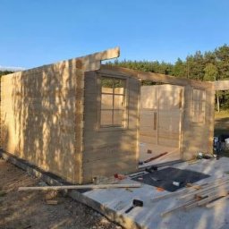 FachBud Rafał Szkwerko - Wznoszenie szkieletu drewnianej altany z widocznymi narzędziami na betonowej podstawie. Częściowo zmontowane ściany z oknami i drzwiami na tle zieleni.