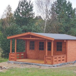 FachBud Rafał Szkwerko - Drewniana altana z tarasem, pomalowana na brązowo, na tle zieleni drzew. Widoczne okna i drzwi. Konstrukcja z bali, szary dach. Otoczenie naturalne, trawa i drzewa.