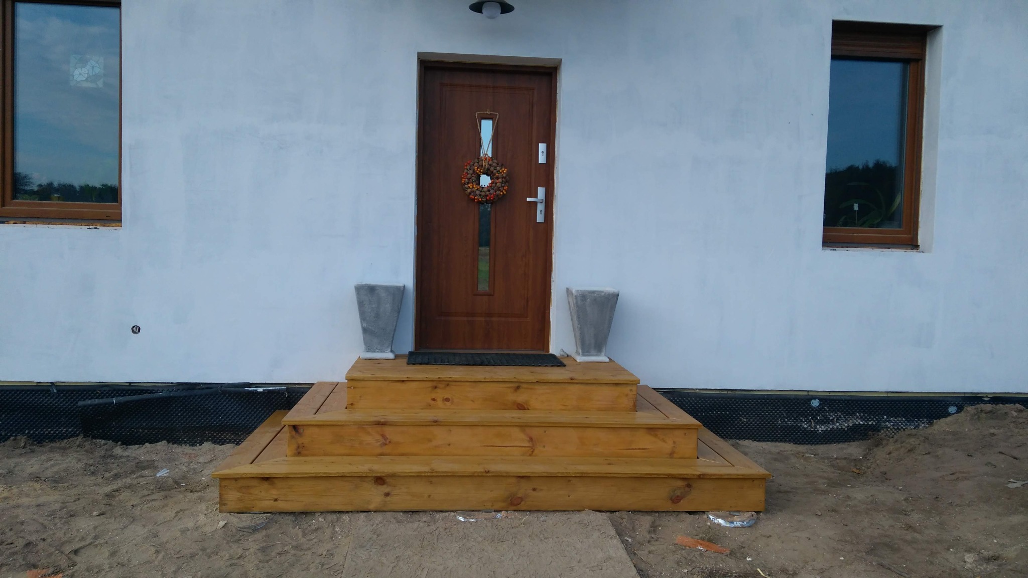 Wejście do domu z drewnianymi schodami i dekoracyjnym wieńcem na brązowych drzwiach. Dwa okna z brązowymi ramami po bokach. Biała elewacja budynku.