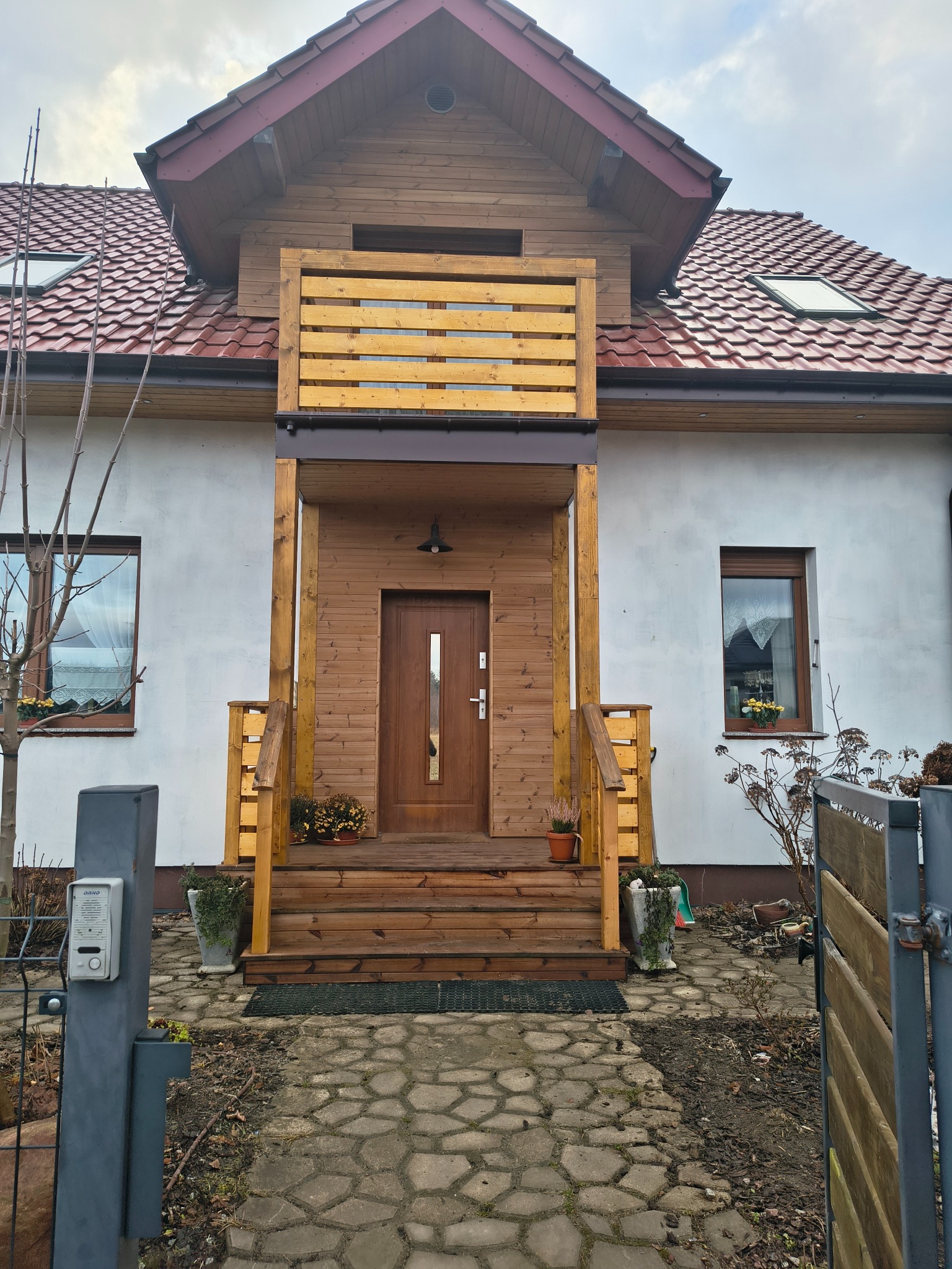 Dwukondygnacyjny dom z drewnianym gankiem i balustradą, widok z zewnątrz. Czerwona dachówka, białe ściany, drewniane wykończenia. Kostka brukowa prowadzi do wejścia.