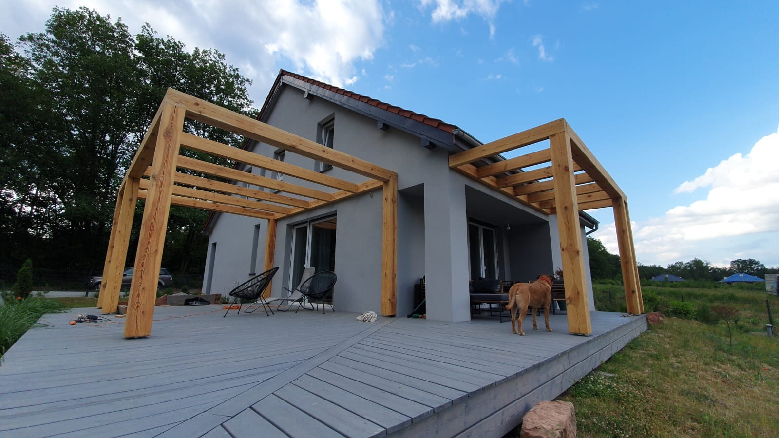 Szary dom z drewnianym tarasem i pergolą w trakcie budowy. Widoczny pies i narzędzia. Ujęcie z dołu, podkreślające konstrukcję i otoczenie.