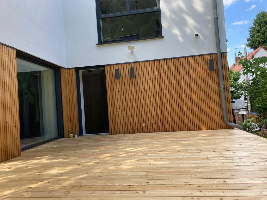 Nowoczesny taras z jasnego drewna, zintegrowany z elewacją budynku wykończoną drewnianymi panelami. Widoczne okno i drzwi tarasowe z czarnymi ramami.