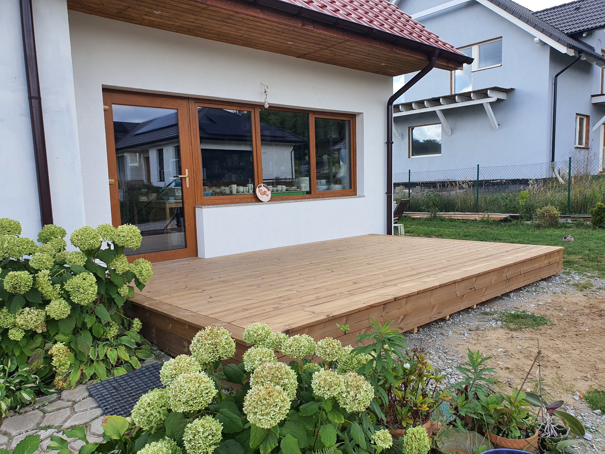 Jasny, drewniany taras przylegający do białego domu z brązowymi oknami, otoczony zielenią i kwiatami hortensji. Widoczny fragment sąsiedniego domu.
