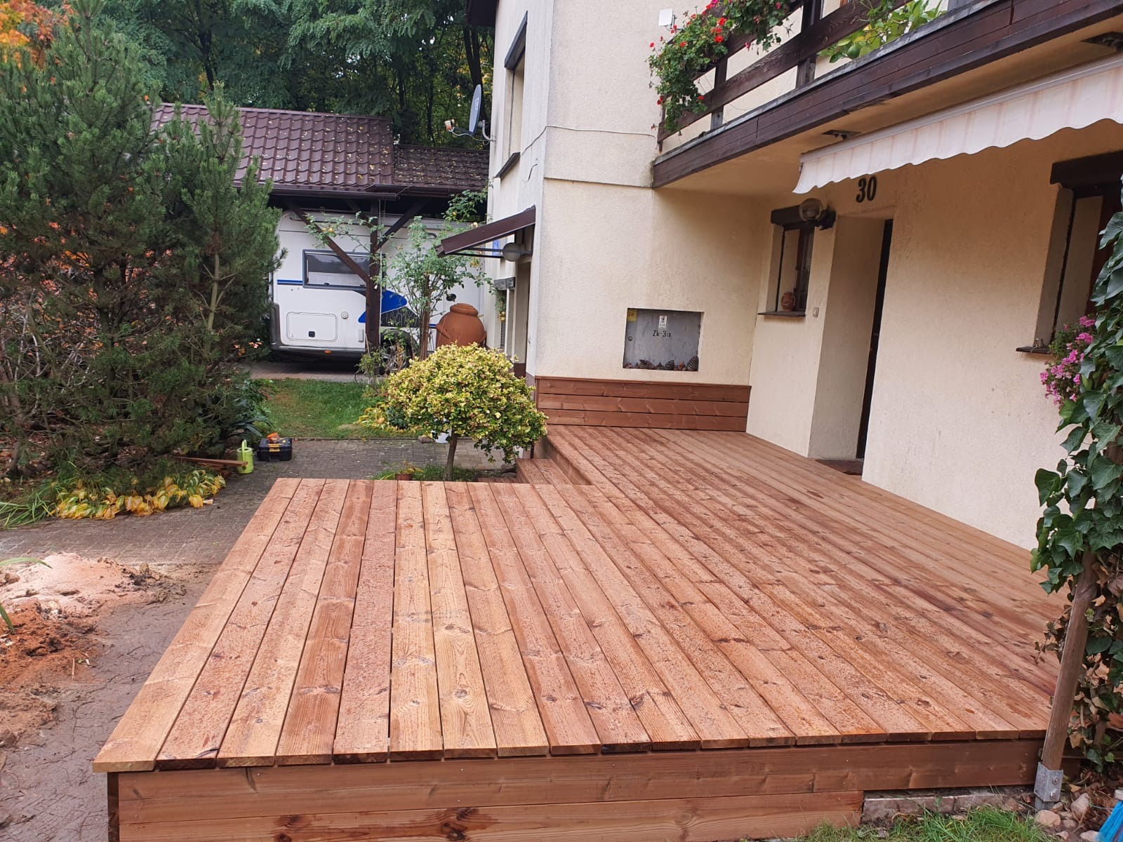 Nowy, drewniany taras przydomowy z desek w ciepłym odcieniu brązu, z niskim stopniem i roślinnością w tle. Widoczny fragment elewacji budynku z numerem 30.