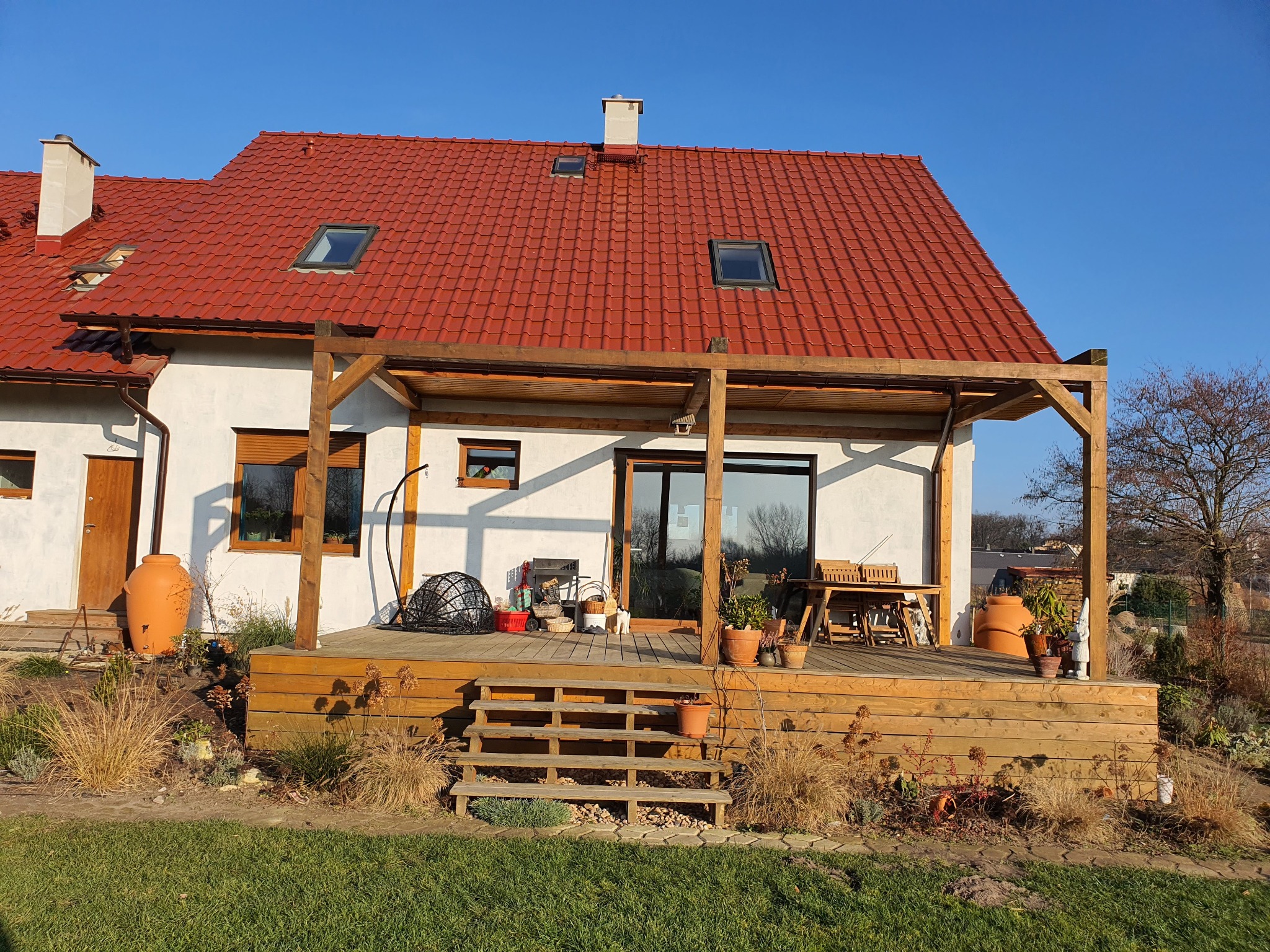 Jasny dom z czerwonym dachem i drewnianym tarasem z pergolą. Na tarasie meble ogrodowe i dekoracje. Uwagę przykuwa duża, gliniana amfora przy ścianie domu.