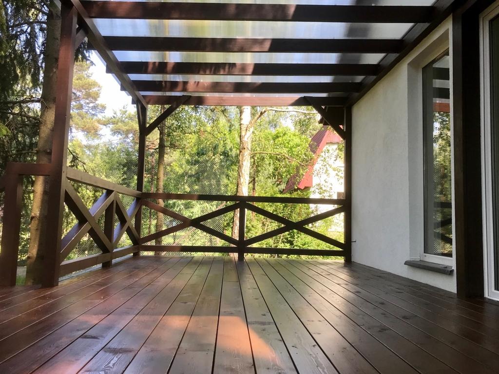 Zadaszony taras drewniany z balustradą krzyżową, świeżo po impregnacji, z widokiem na zieleń. Element domu z oknem po prawej stronie.
