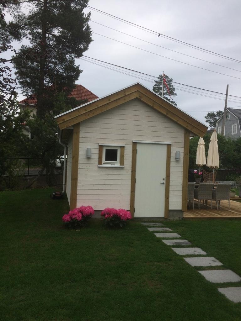 Mała, biała altana z drewnianymi detalami, oświetlona lampami ściennymi, otoczona zielonym trawnikiem i ścieżką z płyt. W tle widoczny taras i meble ogrodowe.