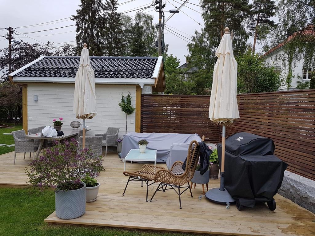 Jasny taras z drewna z meblami ogrodowymi, grillem pod pokrowcem i drewnianą balustradą. W tle widoczny mały budynek z ciemnym dachem i zieleń drzew.