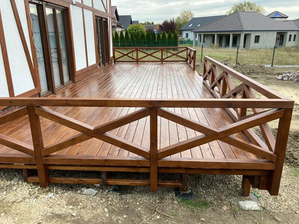 Błyszczący, drewniany taras z balustradą w kolorze orzecha, przylegający do domu z elementami szachownicy na elewacji. W tle widoczny krajobraz z domami.