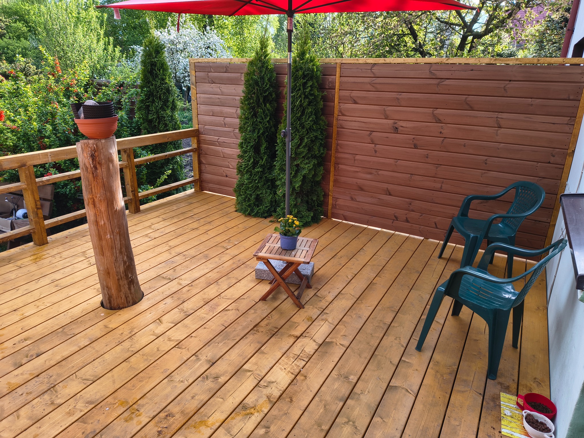 Drewniany taras z brązową deską, zielonymi krzesłami i czerwonym parasolem. Na tarasie rosną tuje, a w tle widać drzewa i krzewy.
