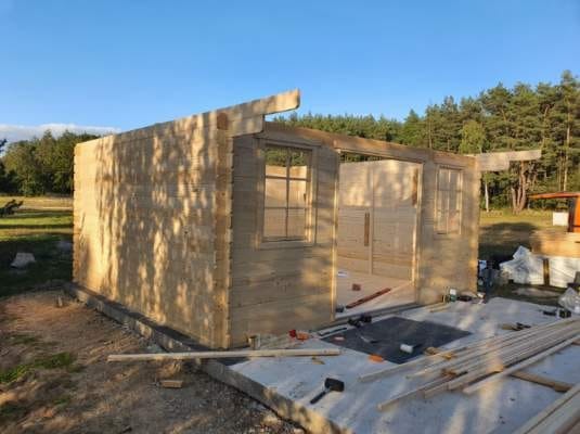 Wznoszenie szkieletu drewnianej altany z widocznymi narzędziami na betonowej podstawie. Częściowo zmontowane ściany z oknami i drzwiami na tle zieleni.