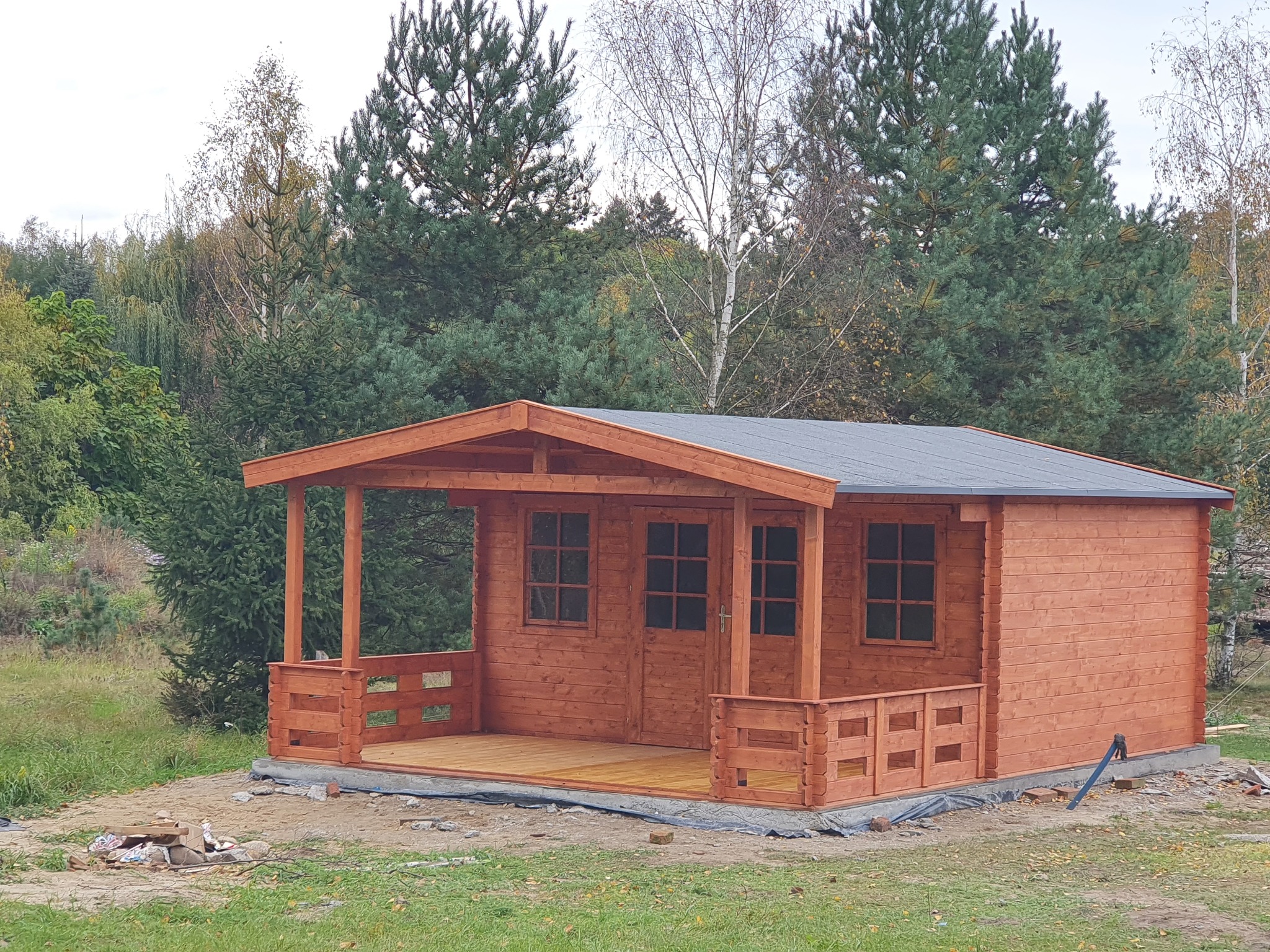 Drewniana altana z tarasem, pomalowana na brązowo, na tle zieleni drzew. Widoczne okna i drzwi. Konstrukcja z bali, szary dach. Otoczenie naturalne, trawa i drzewa.