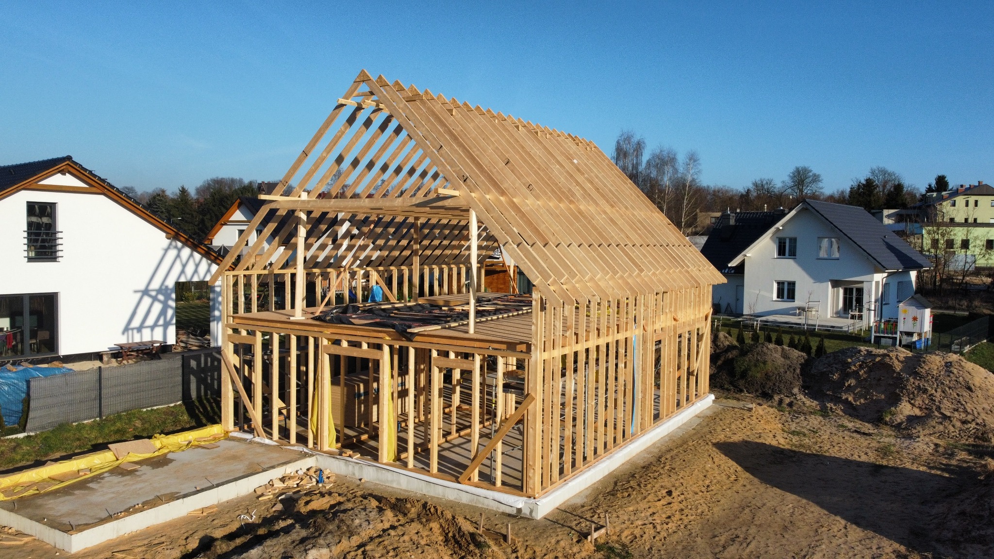 Konstrukcja domu w technologii szkieletowej w trakcie budowy, widok z lotu ptaka, drewniane elementy konstrukcyjne na fundamencie, w tle zabudowa jednorodzinna.