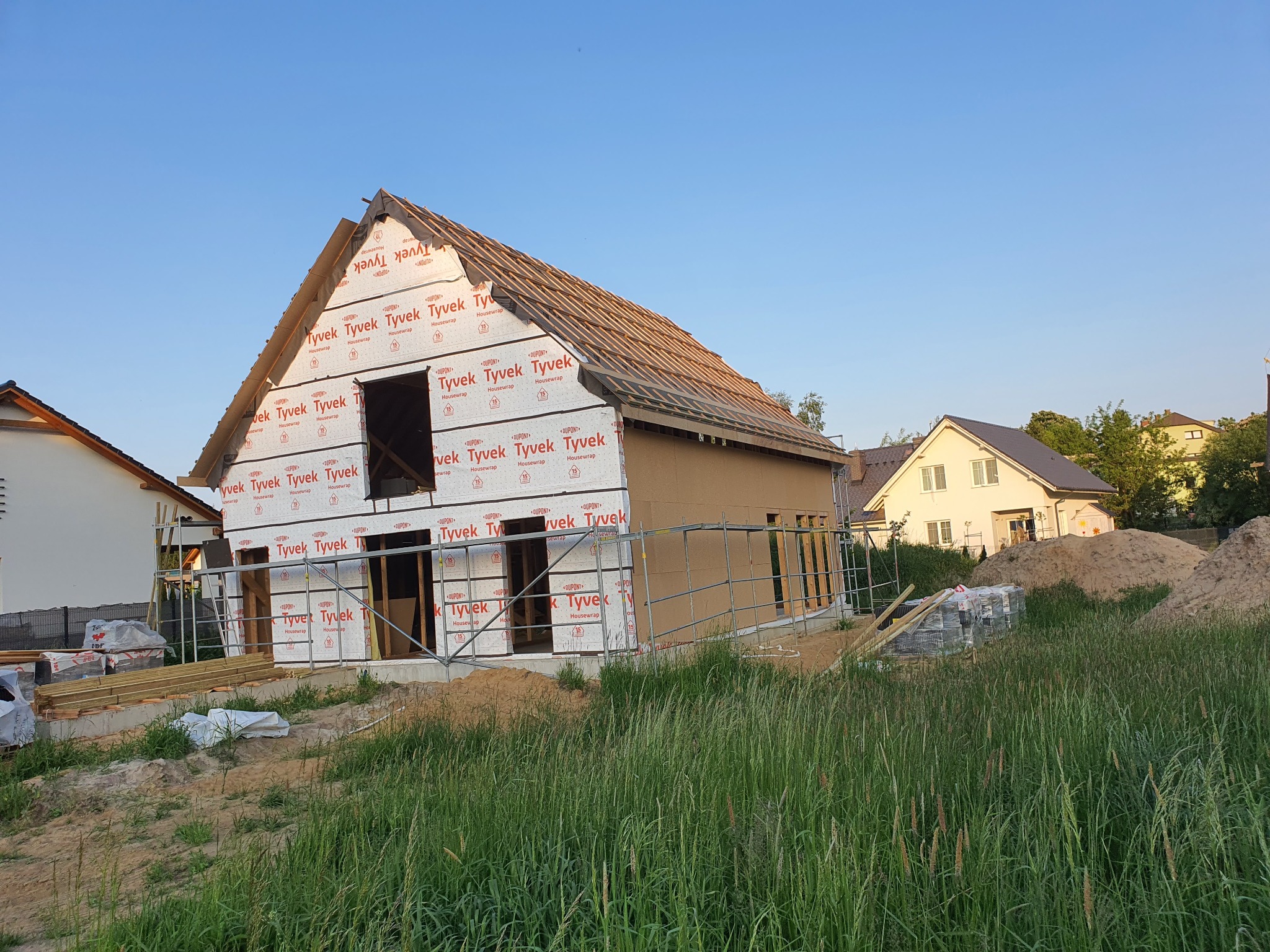 Budowa domu szkieletowego w trakcie: widoczne ściany osłonowe Tyvek, konstrukcja dachu z łatami, rusztowania i materiały budowlane na pierwszym planie.