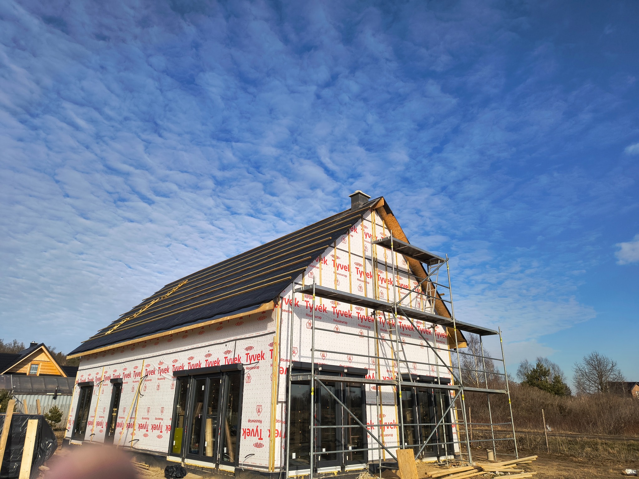 Budowa domu szkieletowego w trakcie: widoczne ściany z membraną Tyvek, rusztowanie i fragment dachu z czarną folią. Błękitne niebo z chmurami w tle.