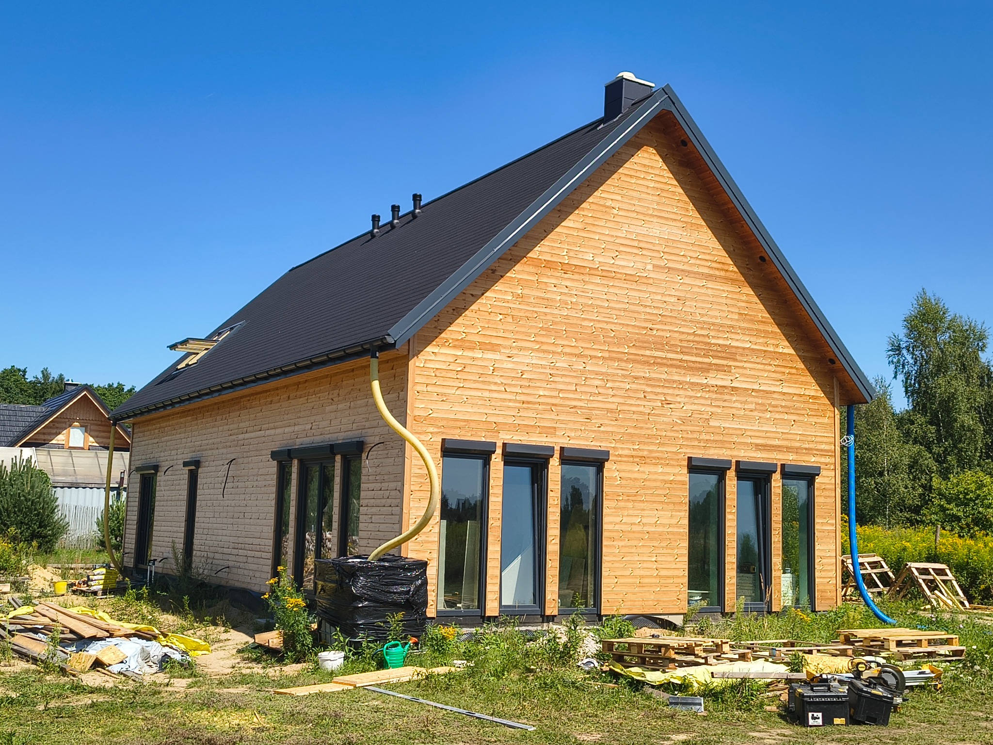 Surowy dom szkieletowy z jasnego drewna pod czarnym dachem, z widocznymi elementami instalacji i narzędziami na zewnątrz, w słoneczny dzień.