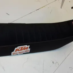 Czarne, odnowione siedzenie motocyklowe KTM z logo Selle Dalla Valle, wykończone pomarańczową nicią, leżące na białym stole roboczym.