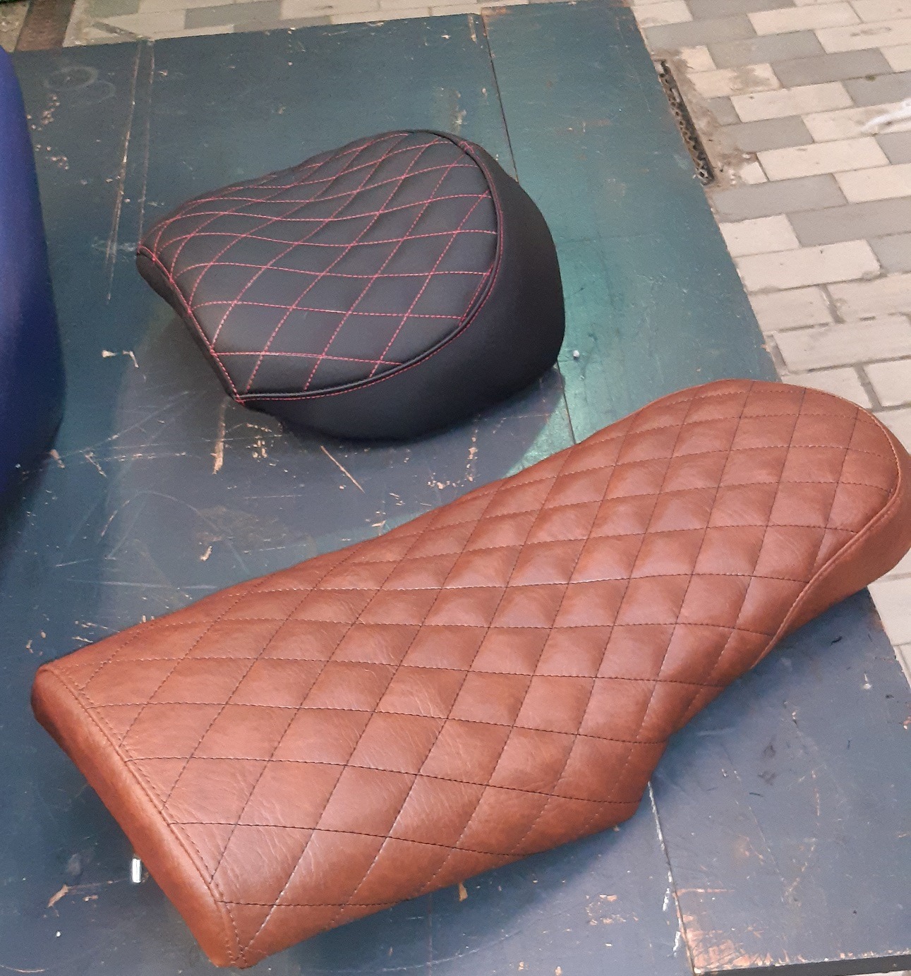 Dwa skórzane siedziska motocyklowe z pikowaniem w romby: brązowe i czarne z czerwonym szwem, leżące na ciemnym, zarysowanym stole.