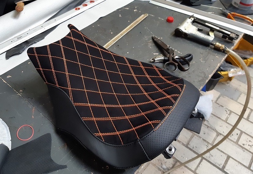 Czarne, pikowane pomarańczową nicią siedzisko motocyklowe z perforowaną skórą na krawędzi, leżące na warsztatowym stole obok narzędzi tapicerskich.