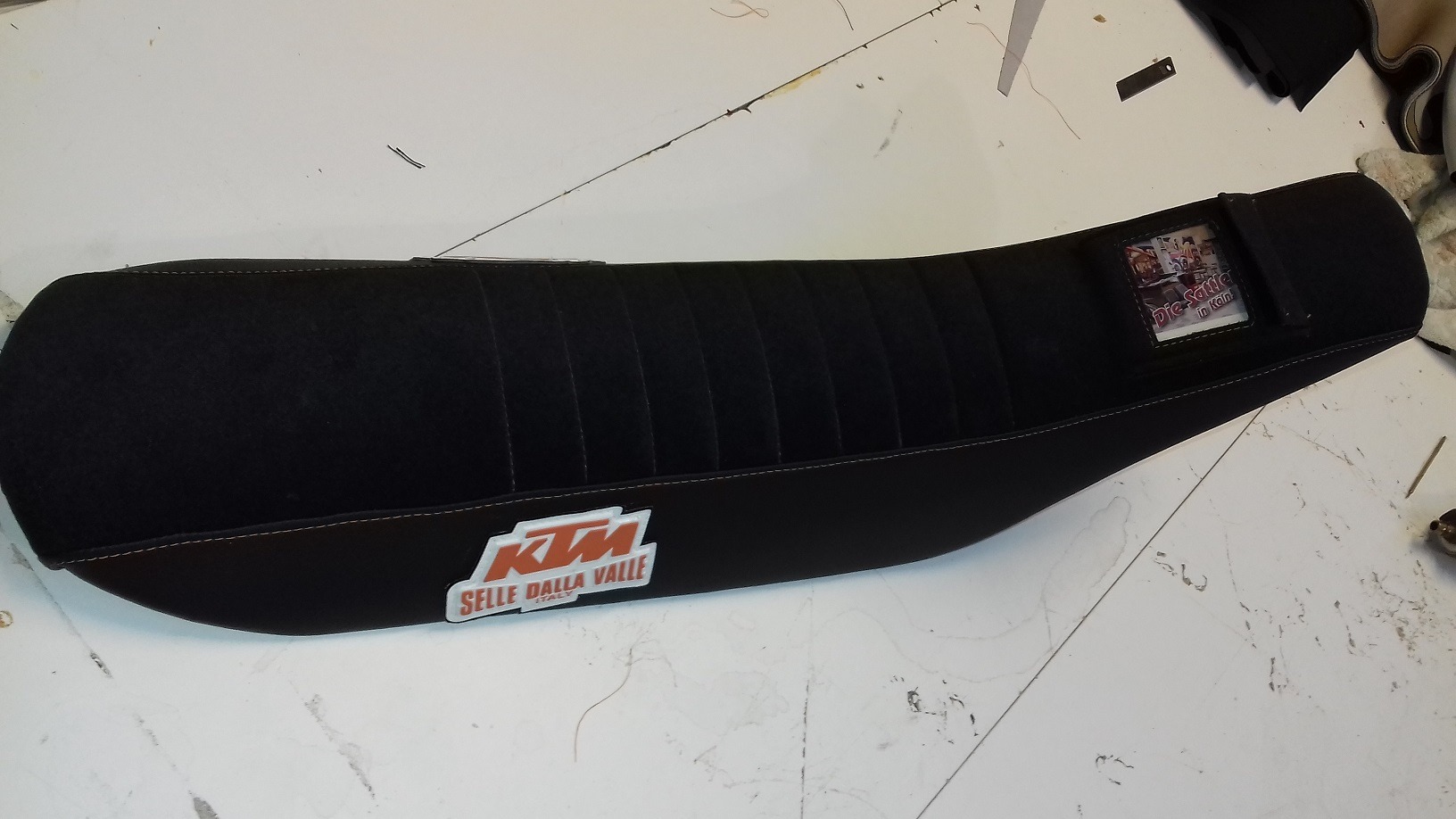 Czarne, odnowione siedzenie motocyklowe KTM z logo Selle Dalla Valle, wykończone pomarańczową nicią, leżące na białym stole roboczym.