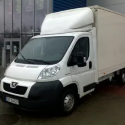 Biały samochód dostawczy Peugeot z zabudową kontenerową, zaparkowany przed przeszklonym budynkiem z niebieską ścianą w tle, widoczny numer rejestracyjny SK 5492P.