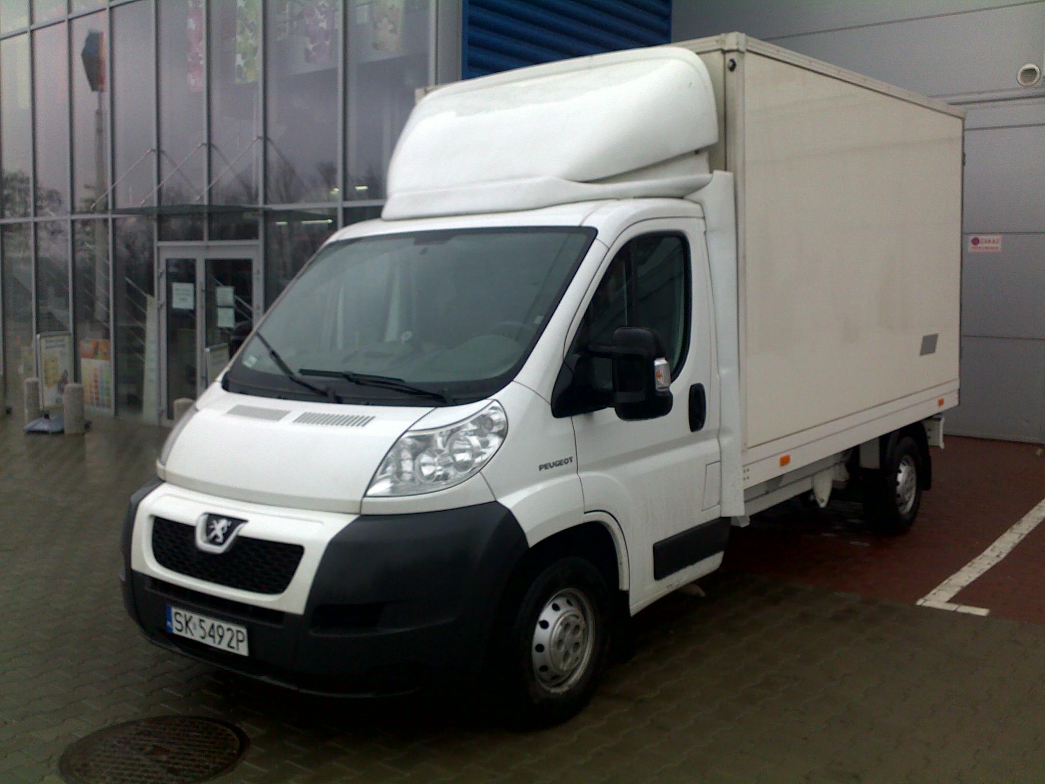 Biały samochód dostawczy Peugeot z zabudową kontenerową, zaparkowany przed przeszklonym budynkiem z niebieską ścianą w tle, widoczny numer rejestracyjny SK 5492P.