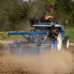 Usługi Agnieszka Ślizak - Niebieski traktor ISEKI z glebogryzarką spulchnia ziemię na polu, za nim zielone drzewa. Operator skupiony na pracy. Przygotowanie terenu pod uprawę.