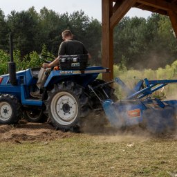 Usługi Agnieszka Ślizak - Niebieski traktor Iseki w trakcie pracy na polu, z widocznym pyłem i świeżo przekopaną ziemią. Operator siedzi na maszynie, w tle las i drewniana konstrukcja.